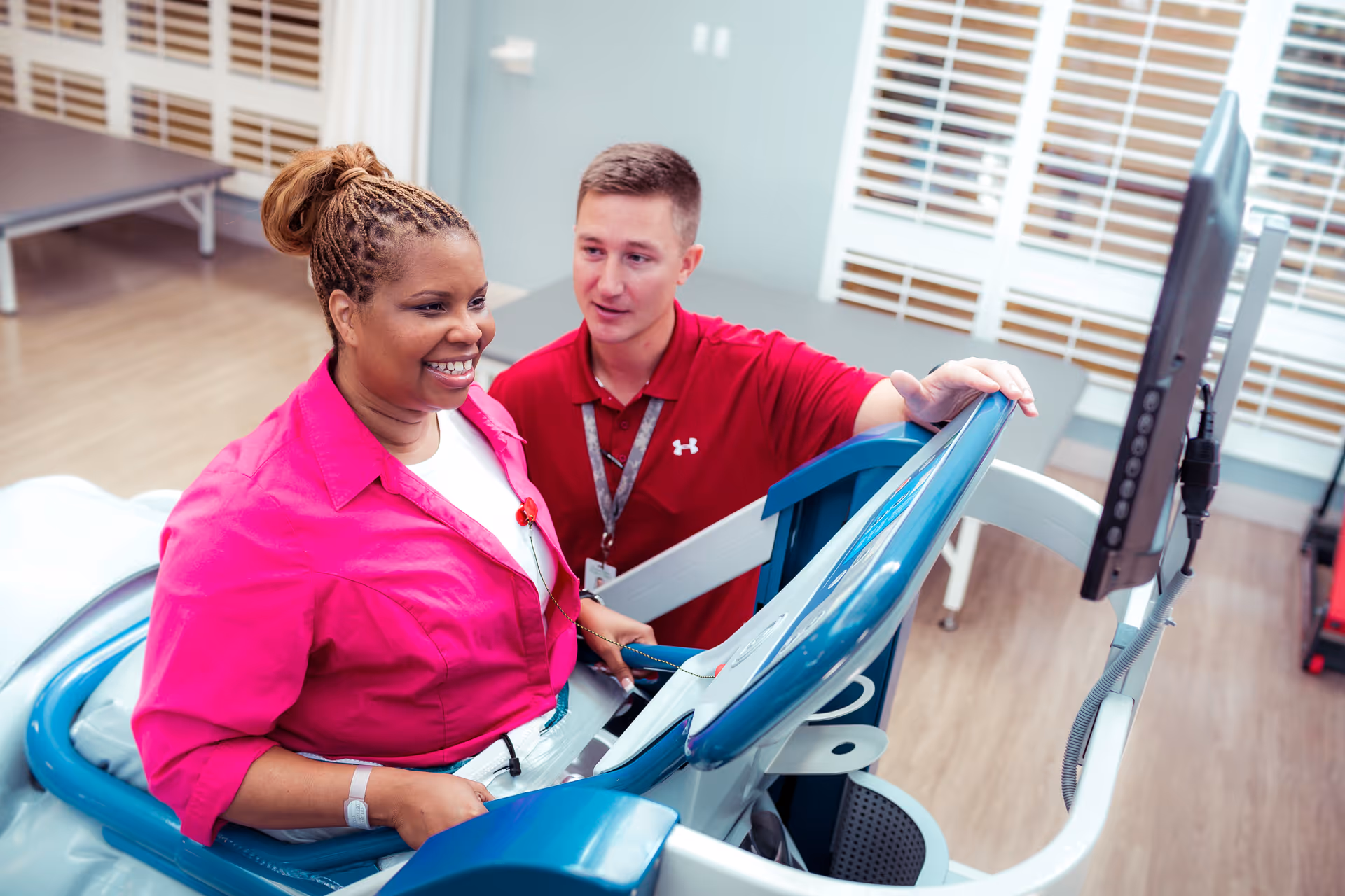 A woman in a bright pink shirt is using a treadmill machine while a man in a red shirt assists her. They are in a room with wooden floors and large windows with white blinds.