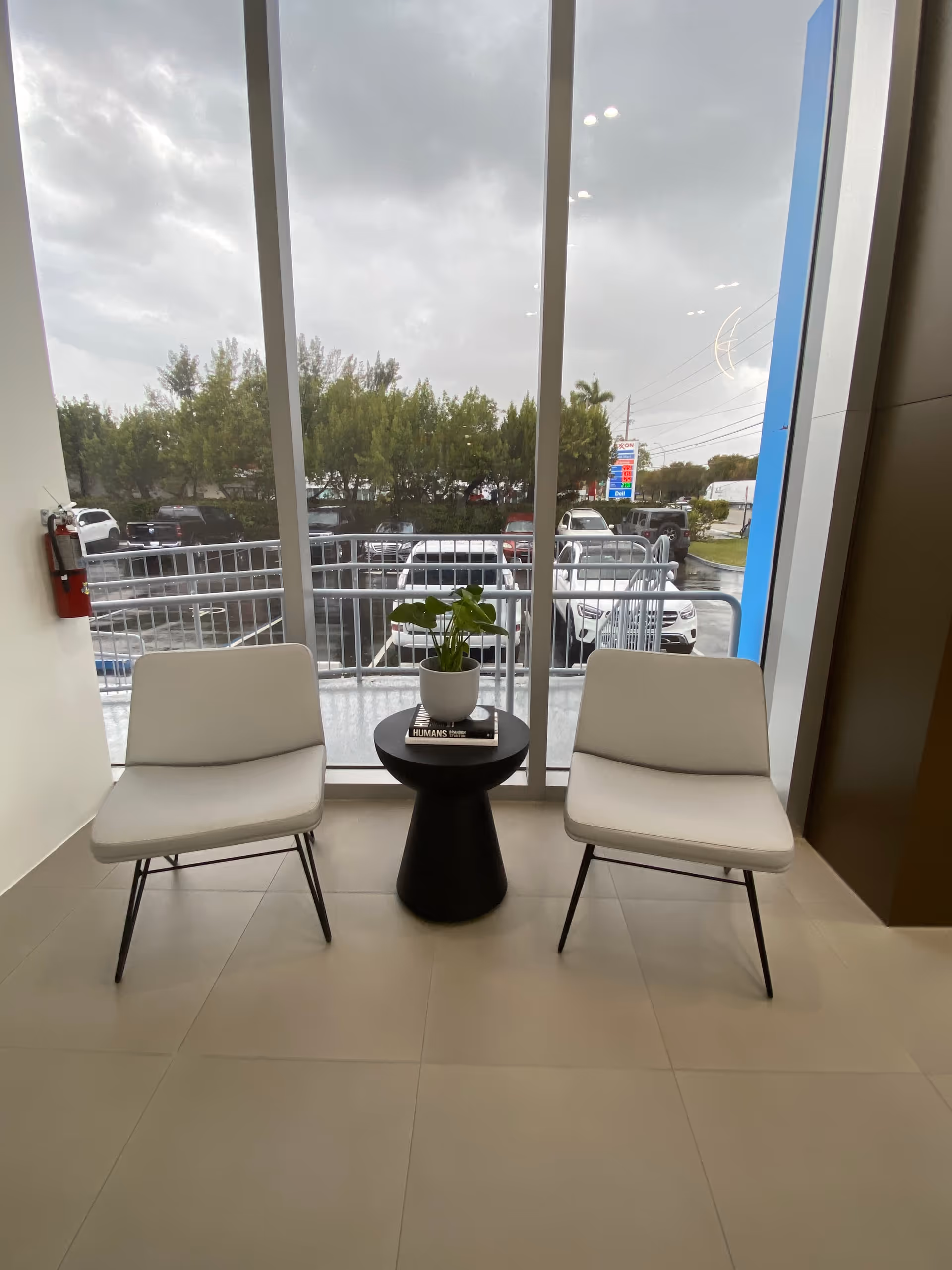 Two white chairs flank a small black table with a plant beside large windows in lobby