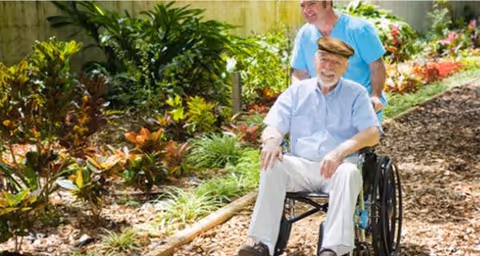An elderly man in a wheelchair outdoors, assisted by a caregiver along a garden path.