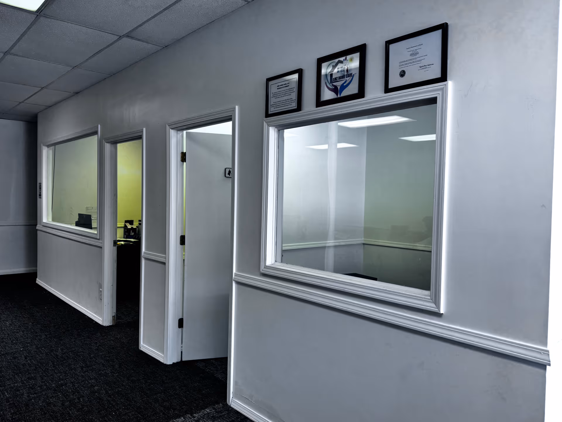 White corridor with office doors, a large windowed wall, and framed certificates above.
