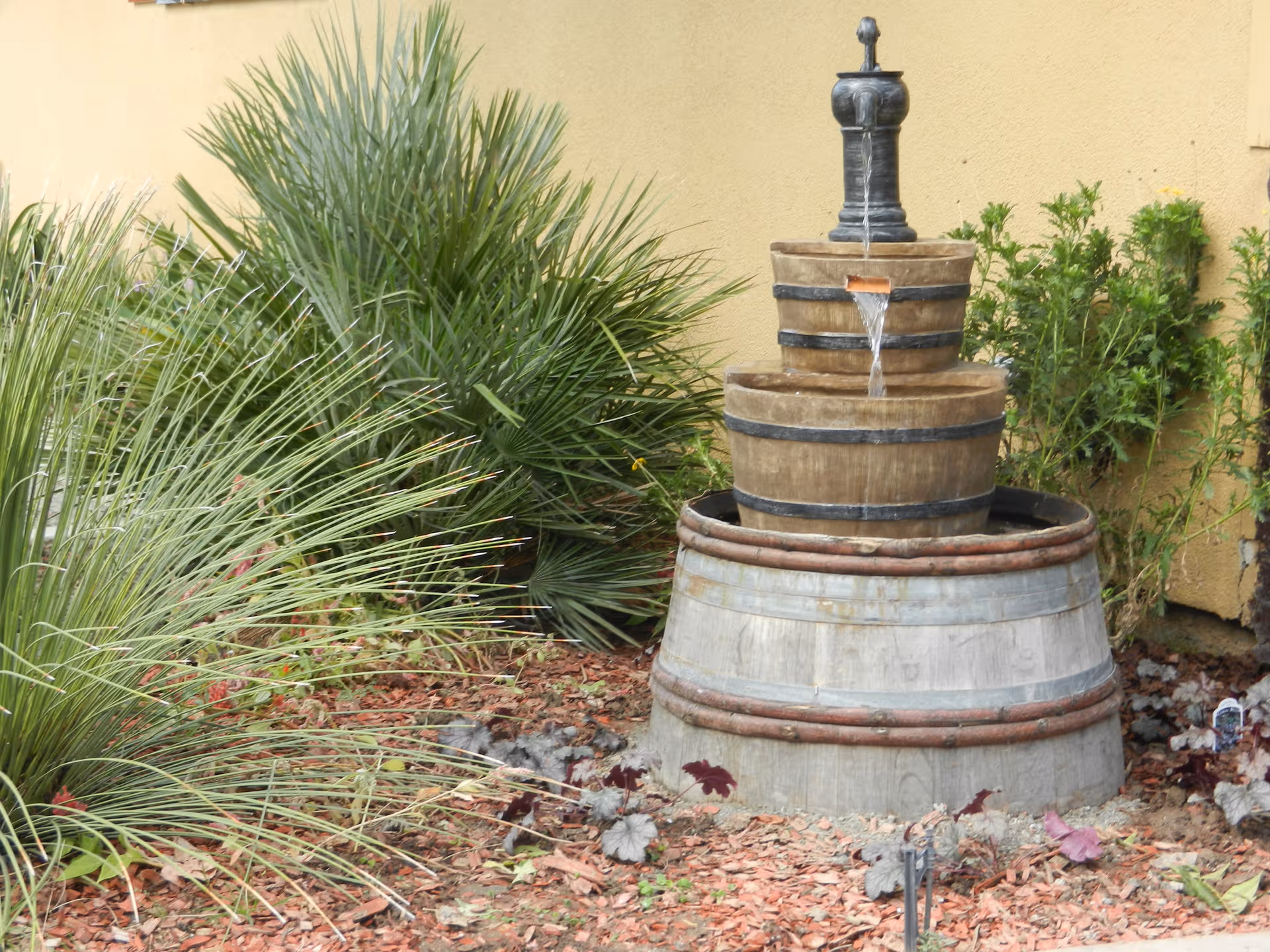Tiered wooden barrel fountain with water spout, surrounded by desert plants beside a beige wall.