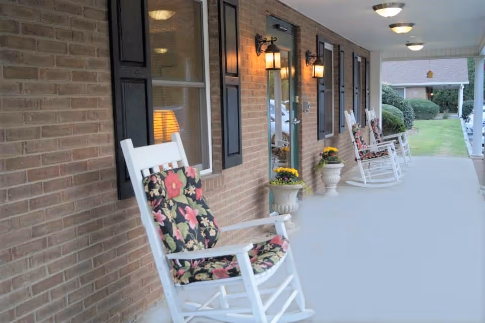 Covered brick porch with white rocking chairs, floral cushions, potted flowers and wall lamps outside an entrance.