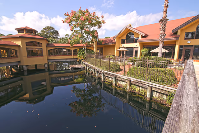 Yellow stucco building with a red roof surrounding a reflective pond, a wooden walkway and landscaped courtyard.