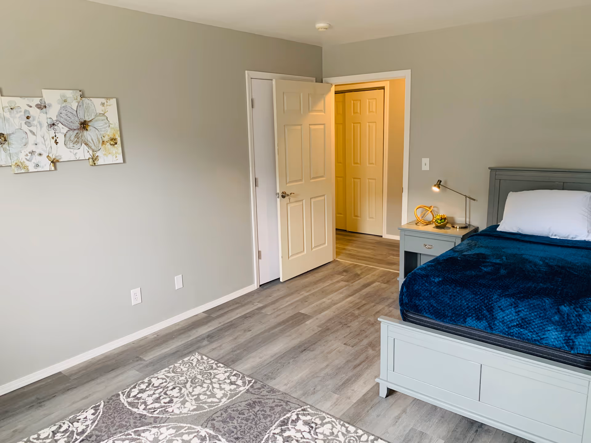 A neatly furnished Hartford Estates bedroom with bed, nightstand, lamp, and doorway to a hallway.