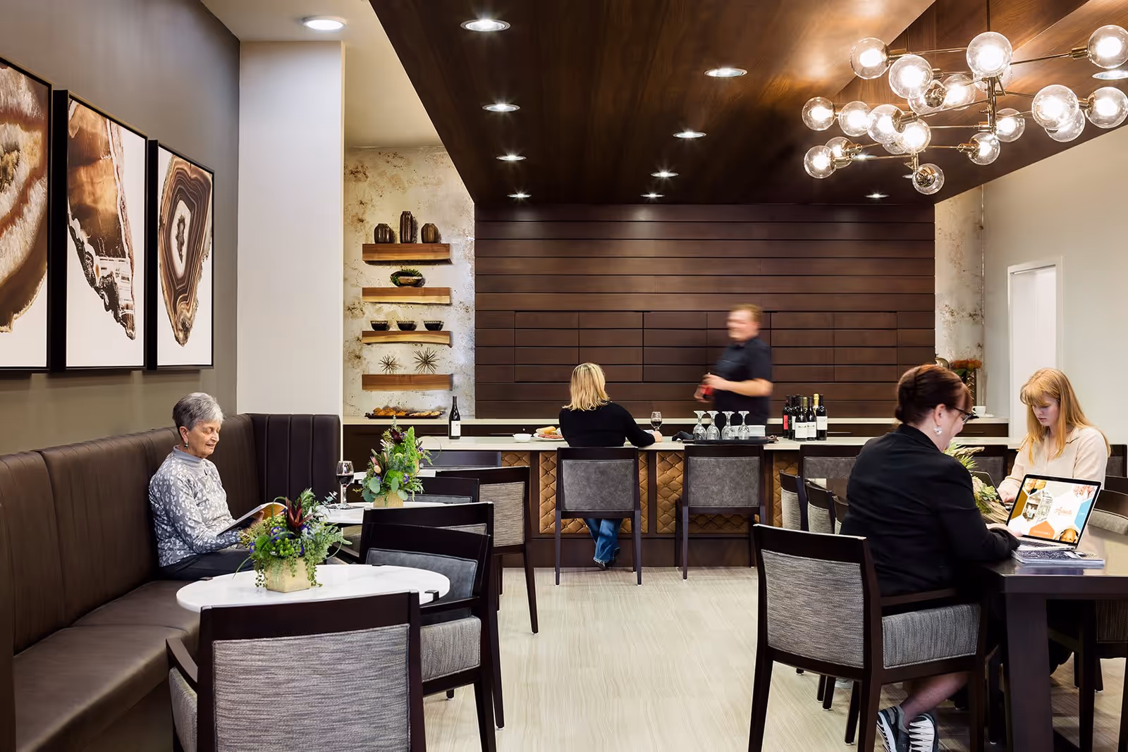 A modern common area with a bar counter and seating. There are four people in the room: an elderly woman sitting alone on a cushioned bench reading a book, a woman sitting at the bar with her back to the camera, a man behind the bar serving drinks, and two women sitting at a table, one working on a laptop. The room features contemporary decor with framed artwork on the wall, plants on tables, and a stylish light fixture hanging from the ceiling.