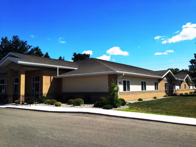 Exterior view of a single-story senior living facility building with a brick and siding facade, surrounded by a paved driveway, small shrubs, and a grassy area under a bright blue sky with scattered clouds.