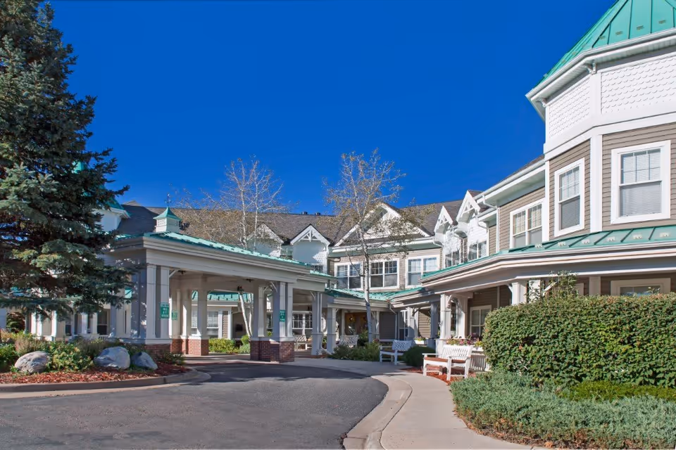 Front entrance and porte-cochère of a multi-story senior living building with green roofs, benches, and landscaped grounds under a clear blue sky.