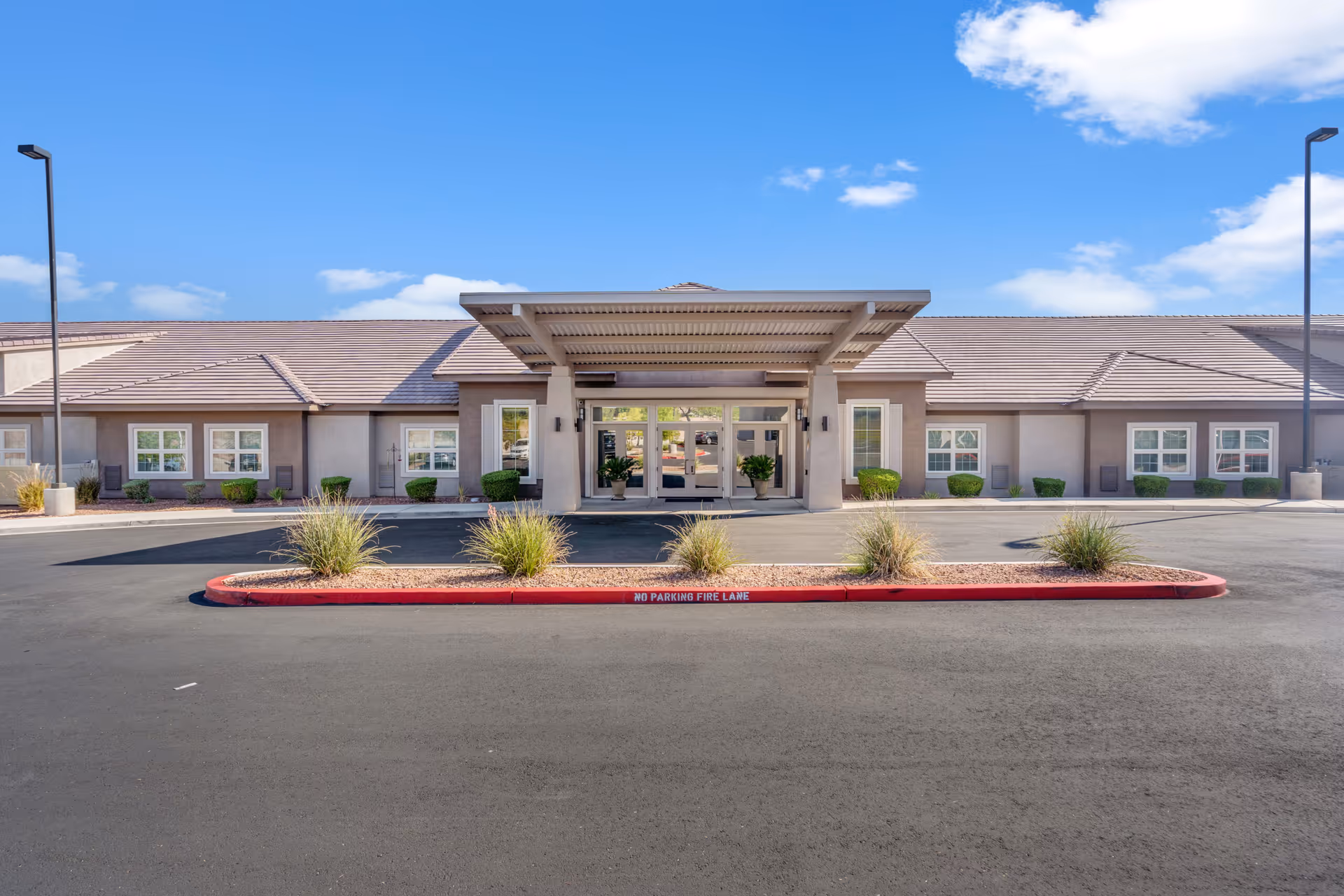 Front exterior view of The Monarch at Henderson facility showing a single-story building with a covered entrance, multiple windows, landscaped bushes, and a clear blue sky.
