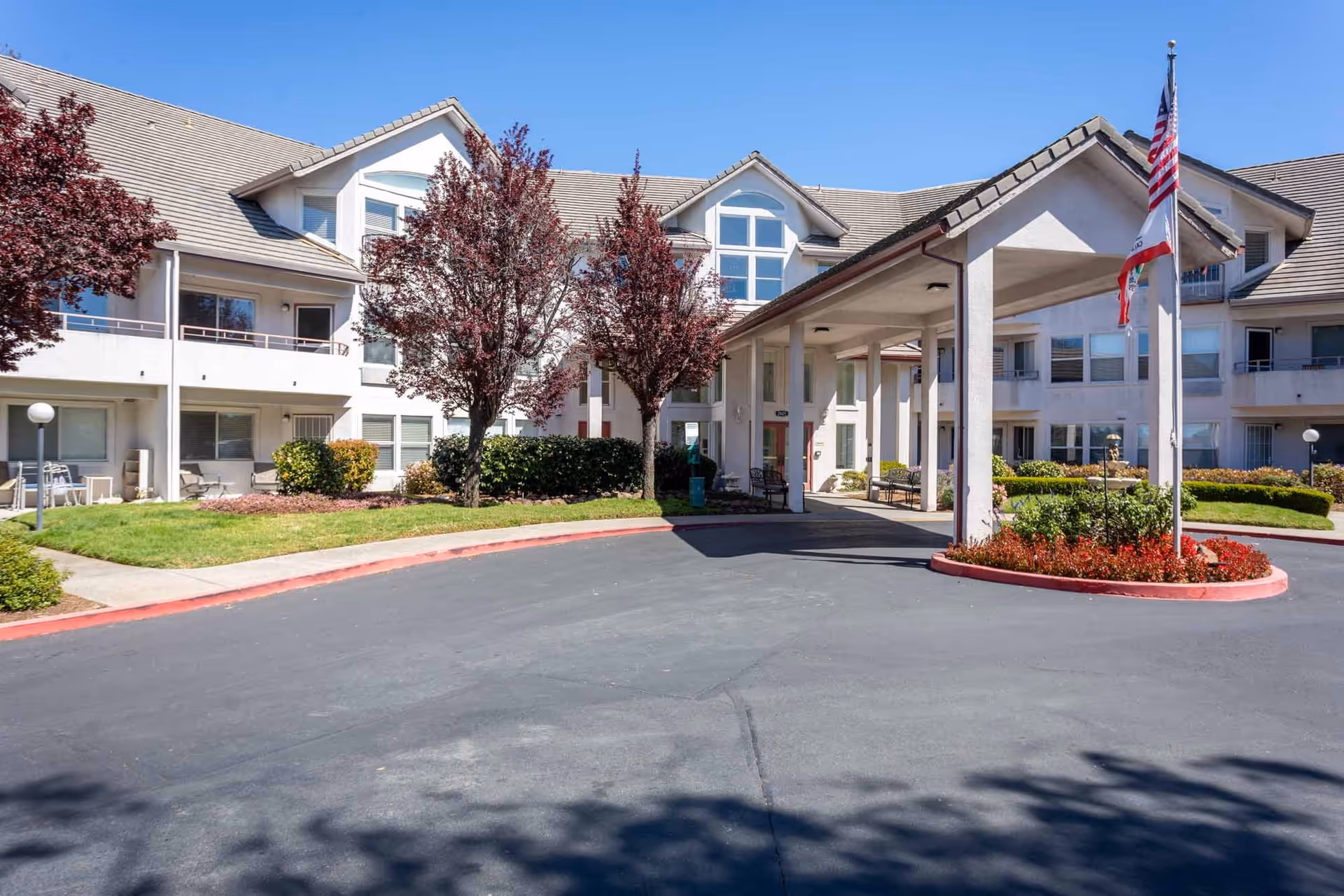 Exterior view of Truewood by Merrill, Pinole senior living facility showing a two-story building with white walls and a covered entrance. There are trees with red leaves, green bushes, and a flagpole with the American and California state flags. The sky is clear and blue.