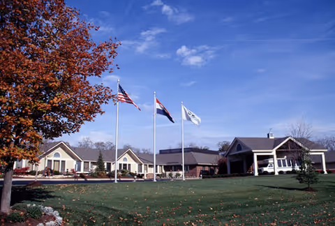 Single-story skilled nursing facility building with three flagpoles on a manicured lawn under a blue sky.