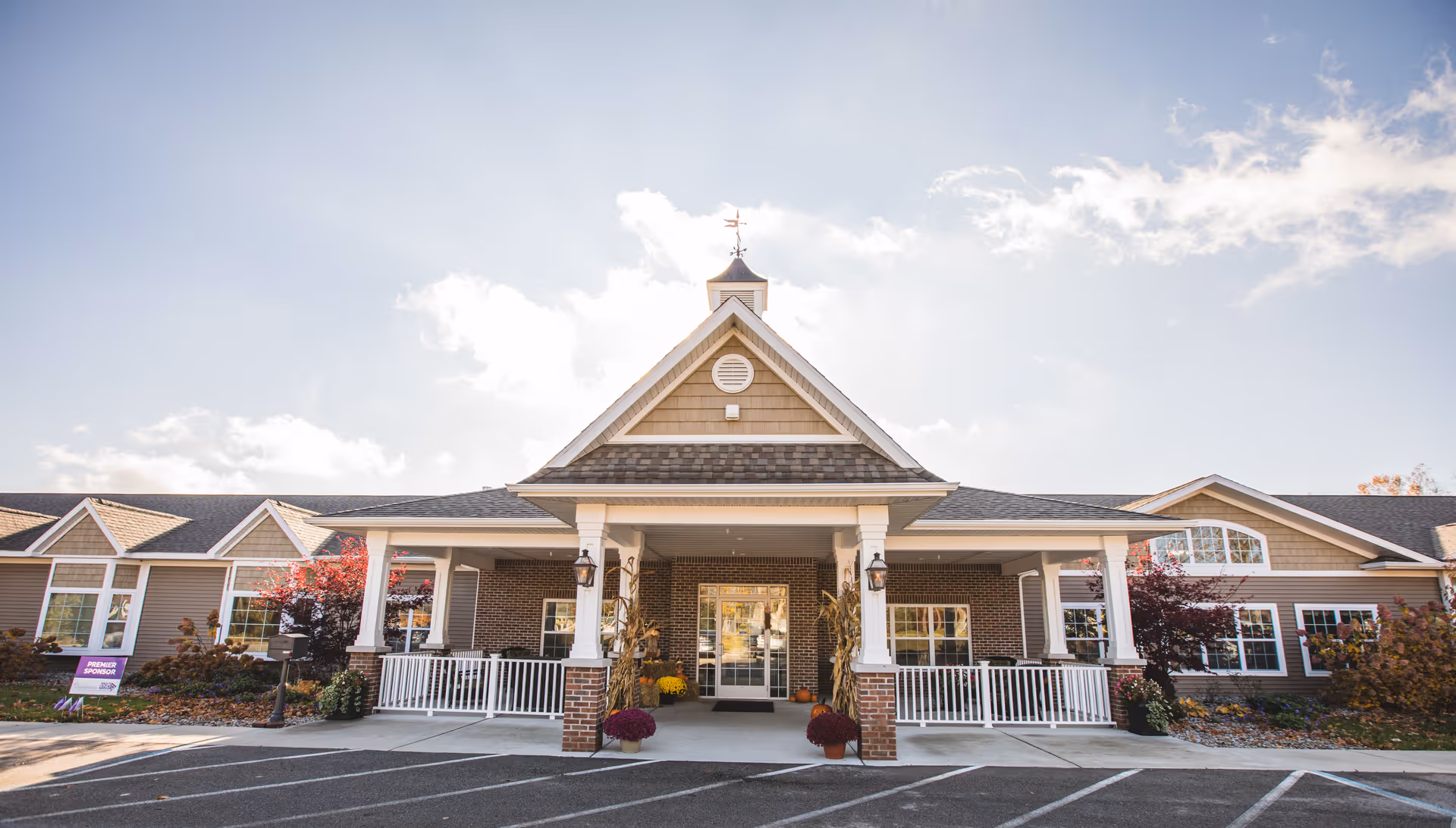 Front exterior view of Gaslight Village Assisted Living & Memory Care building with a covered entrance, white railings, brick pillars, and a clear sky with some clouds.
