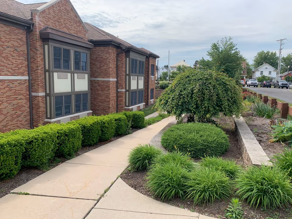 Brick senior living facility exterior with walkway and trimmed shrubs.