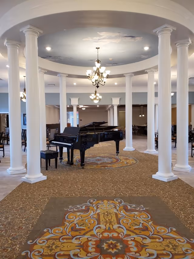 A spacious senior living common room with a grand piano centered under a domed ceiling, surrounded by white columns and chandeliers.