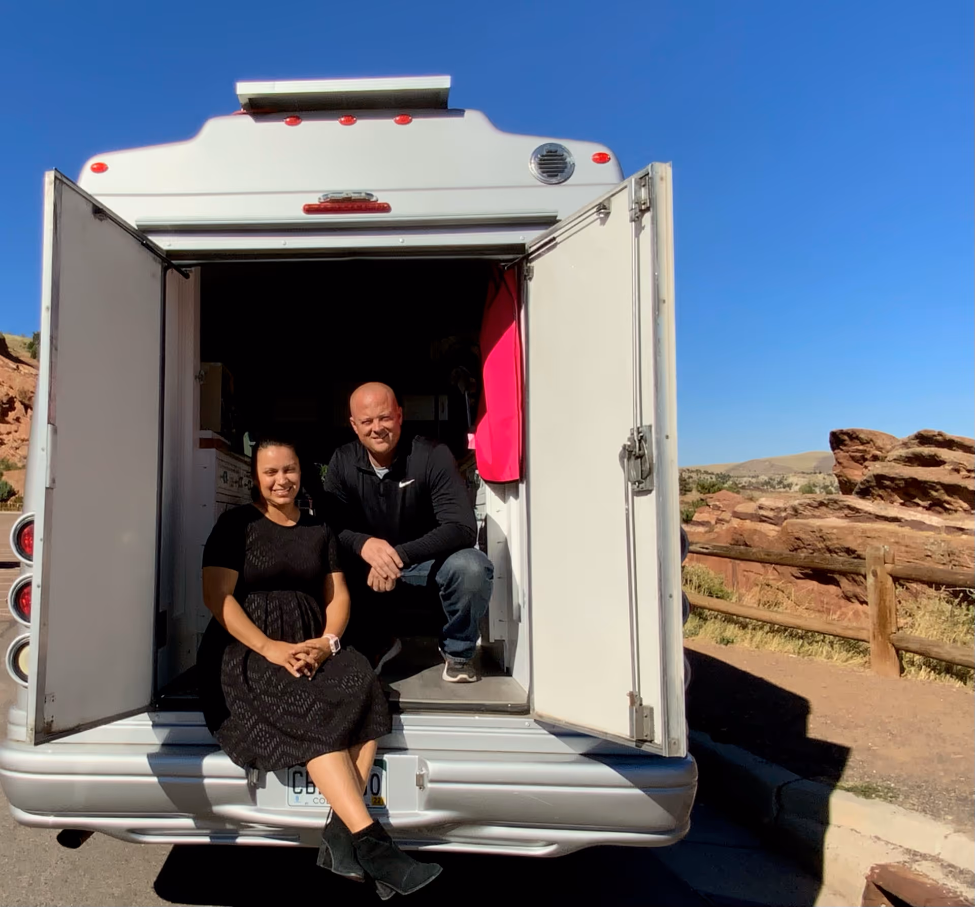 Two people sit at the open rear of a white mobile unit with desert landscape behind.