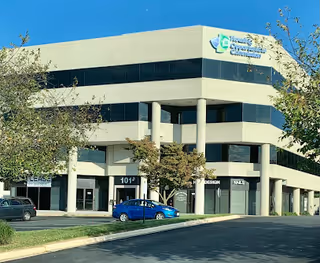 Exterior of a modern, multi-story care facility with glass windows, signage, and a parked blue car.