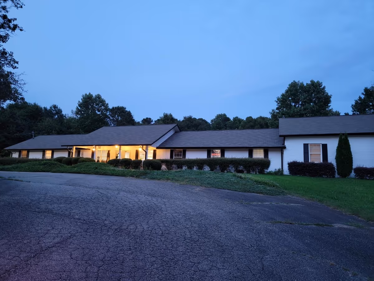 Single-story senior living facility exterior at dusk with lit entrance and landscaped shrubs.