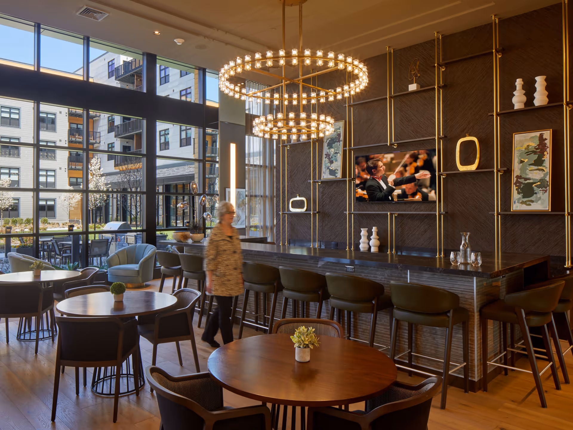 Communal dining and lounge area with round tables, a long bar with stools, large circular chandeliers, floor-to-ceiling windows, and a person walking.