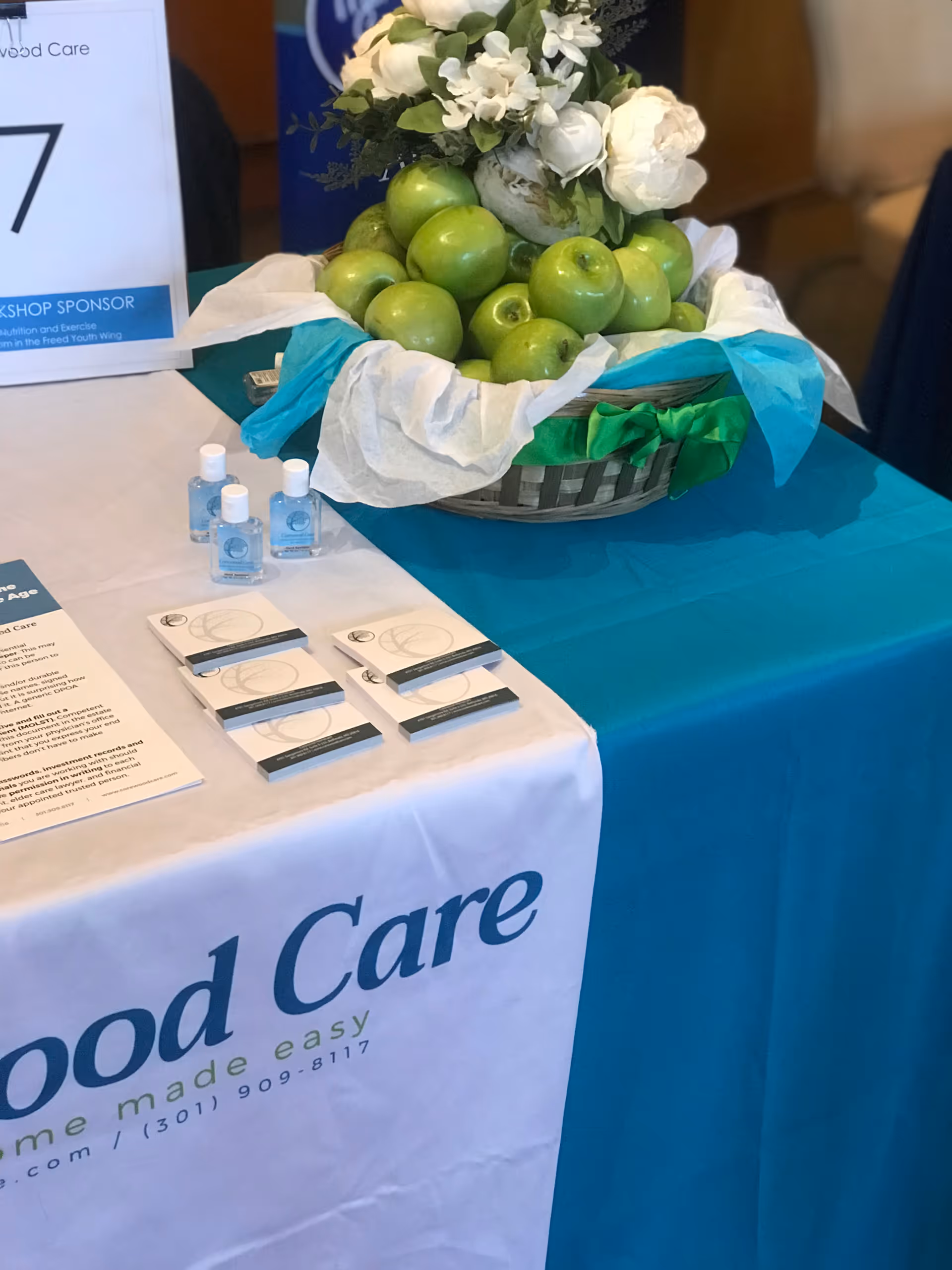 Table display at Corewood Care booth with apple basket, flowers, and brochures.