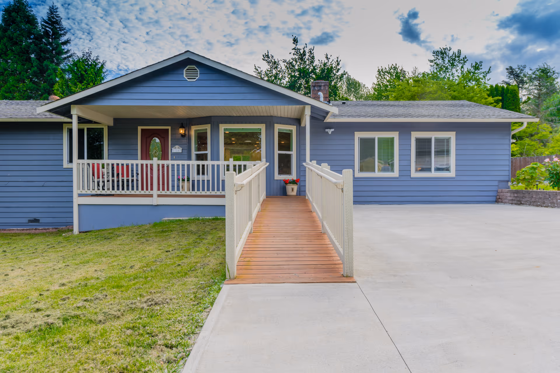 Front exterior view of a single-story blue house with a covered porch and a wooden ramp leading to the front door. The house has white trim around the windows and porch railing, a concrete driveway, and a grassy lawn on the left side. Trees and greenery are visible in the background under a partly cloudy sky.