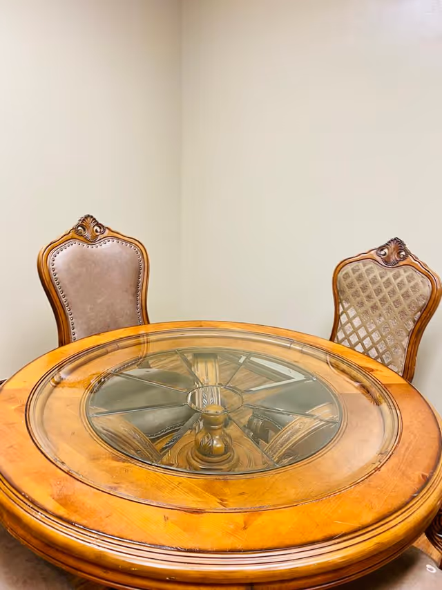 Round wooden dining table with glass top and two ornate chairs in a beige room.