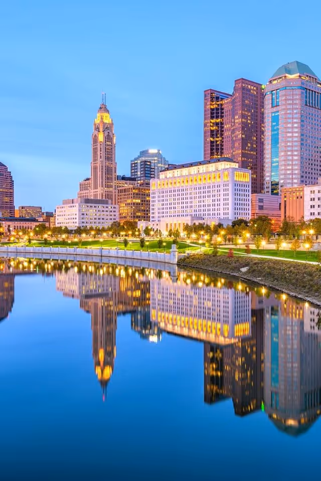 Evening city skyline with illuminated buildings reflecting on a calm river.