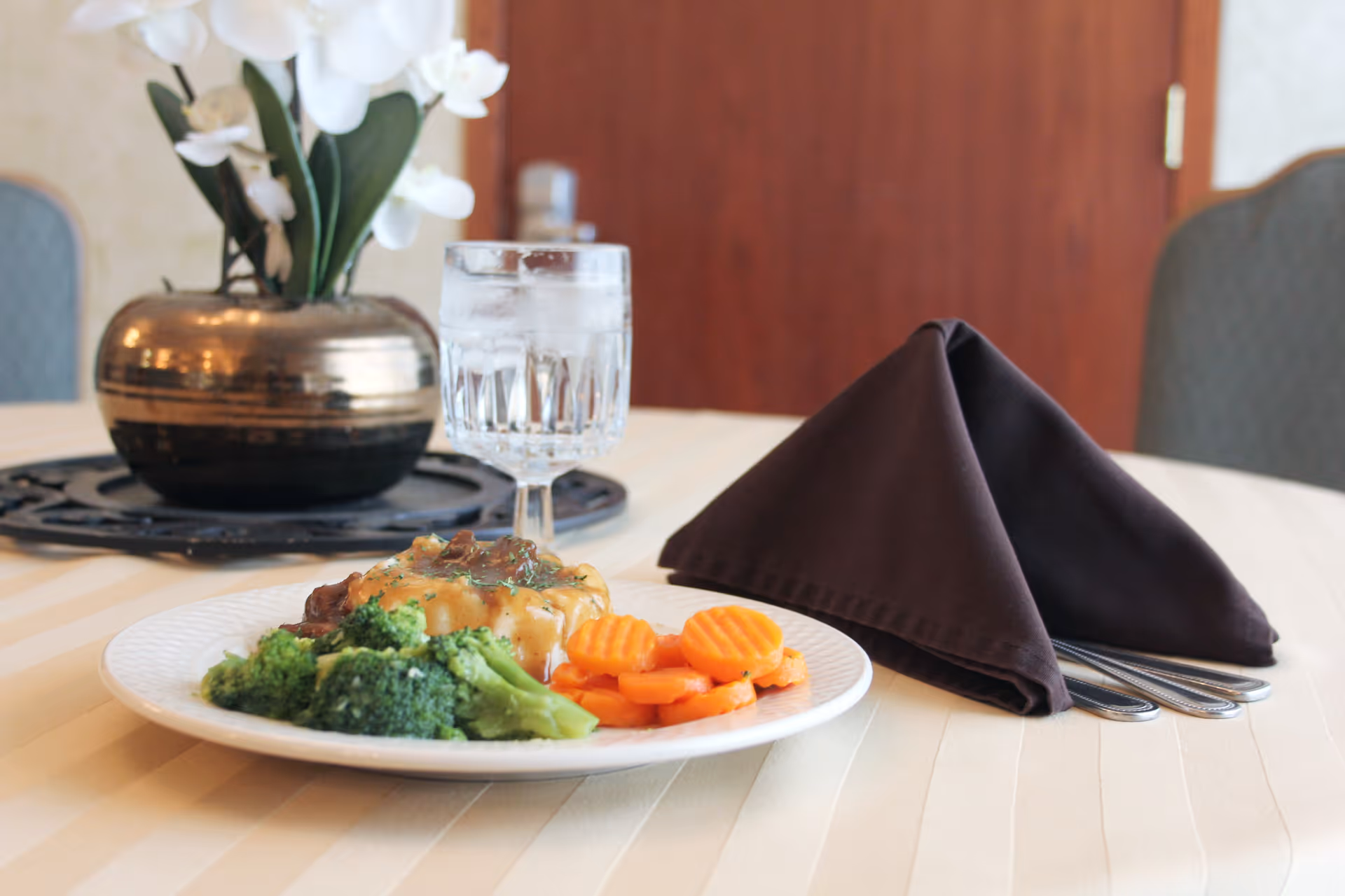 A plated meal of broccoli, carrots, and a meat entrée with a glass of water and a folded napkin on a dining table with a floral centerpiece.