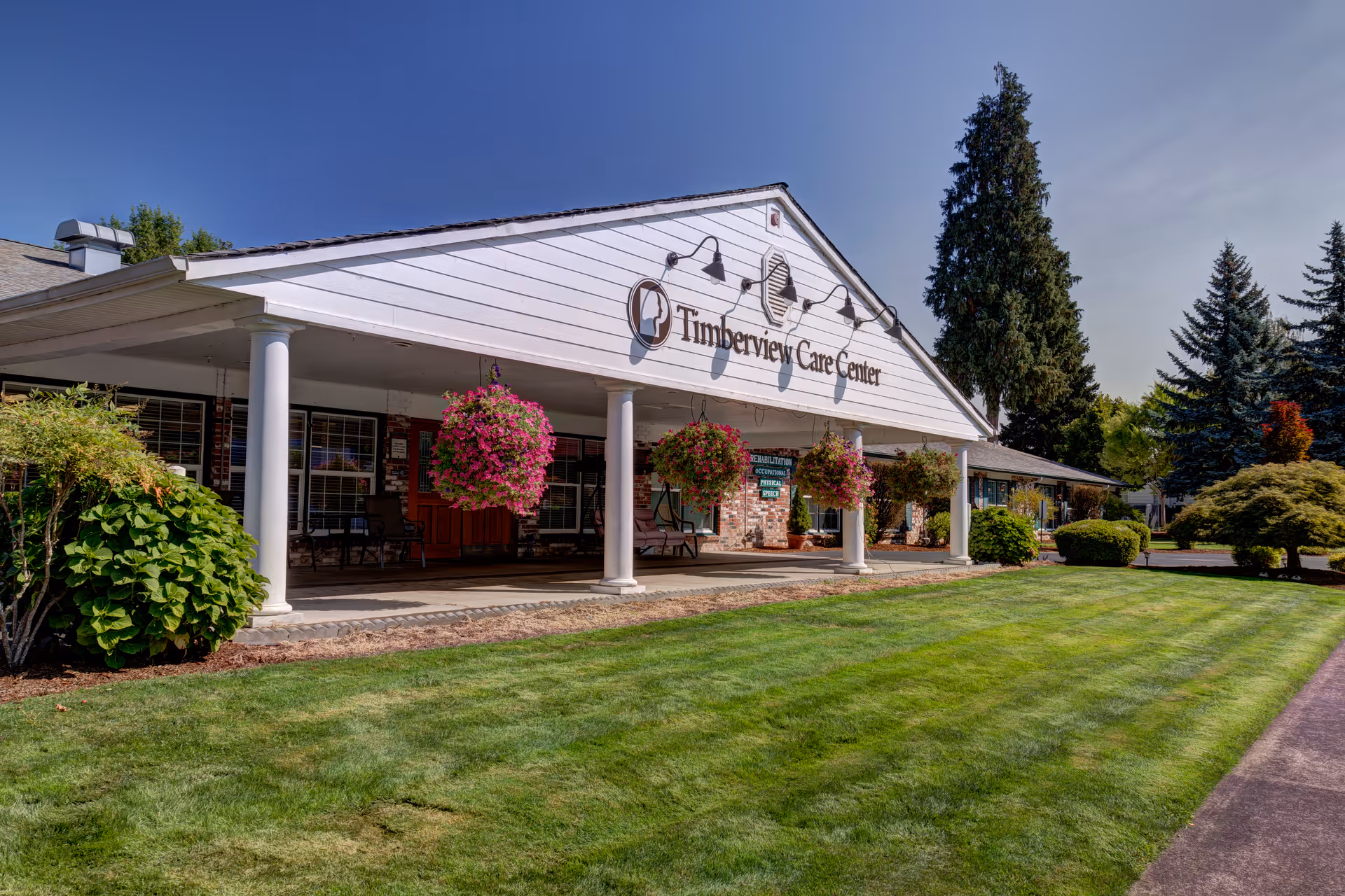 Exterior view of Timberview Care Center building with a covered entrance supported by white columns, hanging flower baskets, well-maintained green lawn, and tall trees in the background under a clear blue sky.