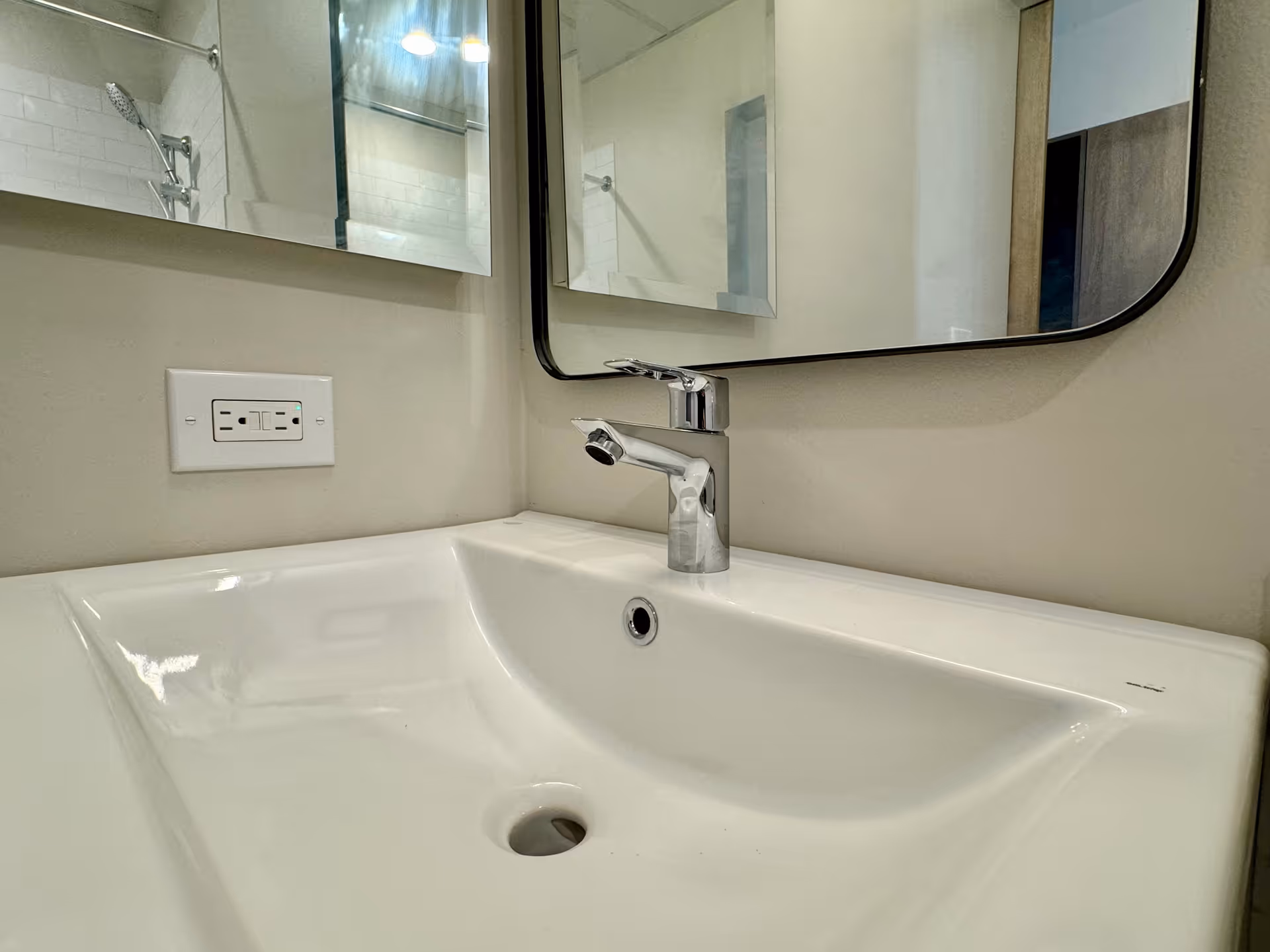 White bathroom sink with chrome faucet and mirror, with a shower area visible in the background.