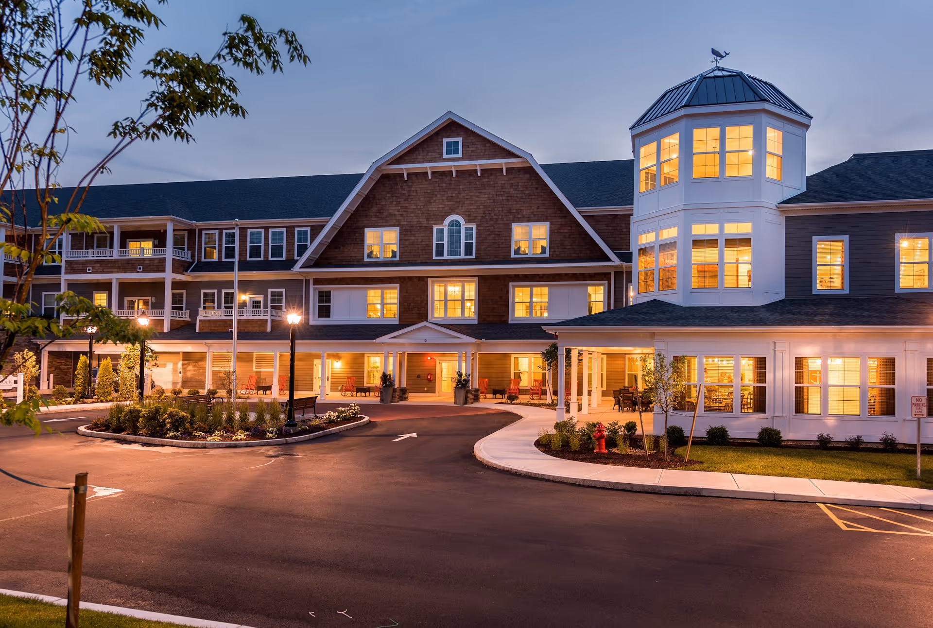 Exterior front of a well-lit, multi-story senior living building with a circular driveway and illuminated windows at dusk.