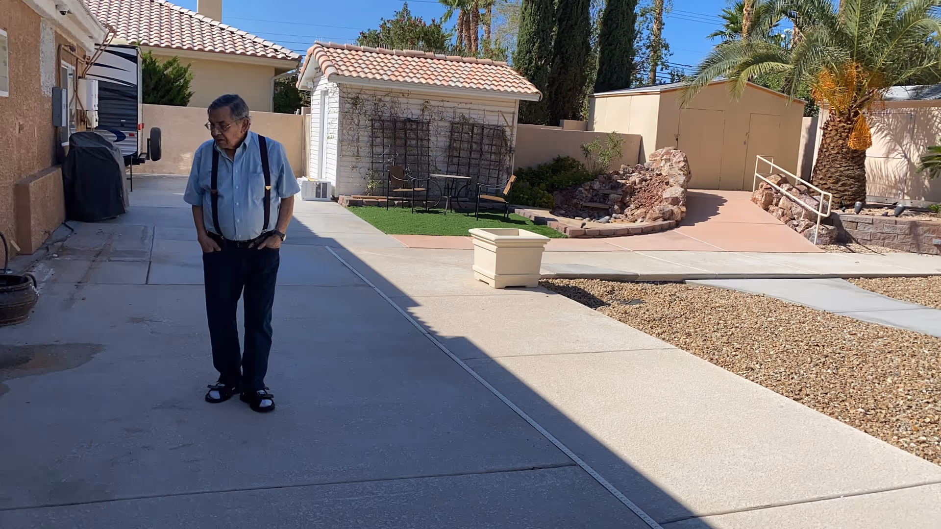 An elderly man stands on a walkway outside a senior residence, with palm trees nearby.