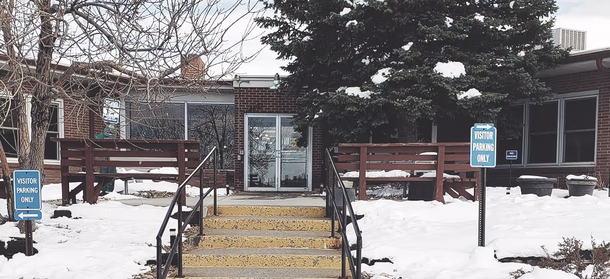 Snow-covered front entrance of a brick care facility with stairs, handrails, benches, and 'Visitor Parking Only' signs.