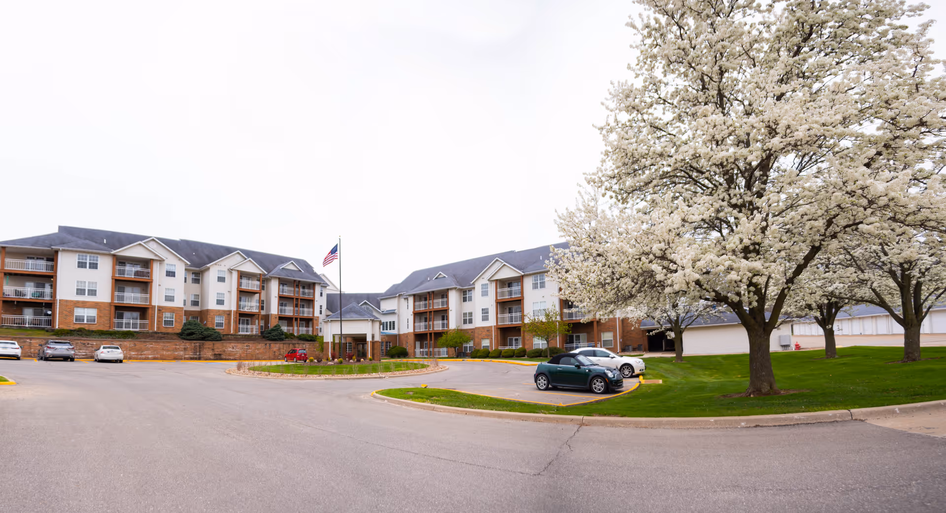 Exterior view of a senior living facility with a curved driveway, parked cars, and blossoming trees.