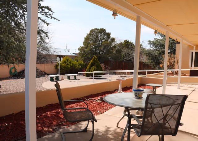 Covered outdoor patio area with a round glass table and three metal chairs. The patio overlooks a landscaped garden with mulch, bushes, trees, and a stone bench. The sky is clear and the area is fenced with a low wall and wooden fence.