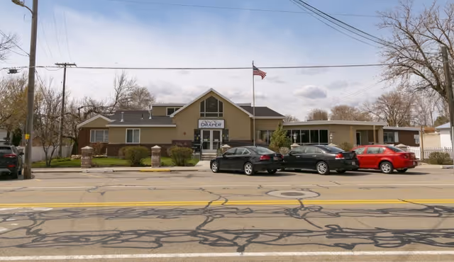 Front exterior view of Draper Rehabilitation and Care Center building with an American flag flying on a pole in front. Several cars are parked in the parking lot in front of the building. Trees without leaves are visible around the property under a partly cloudy sky.