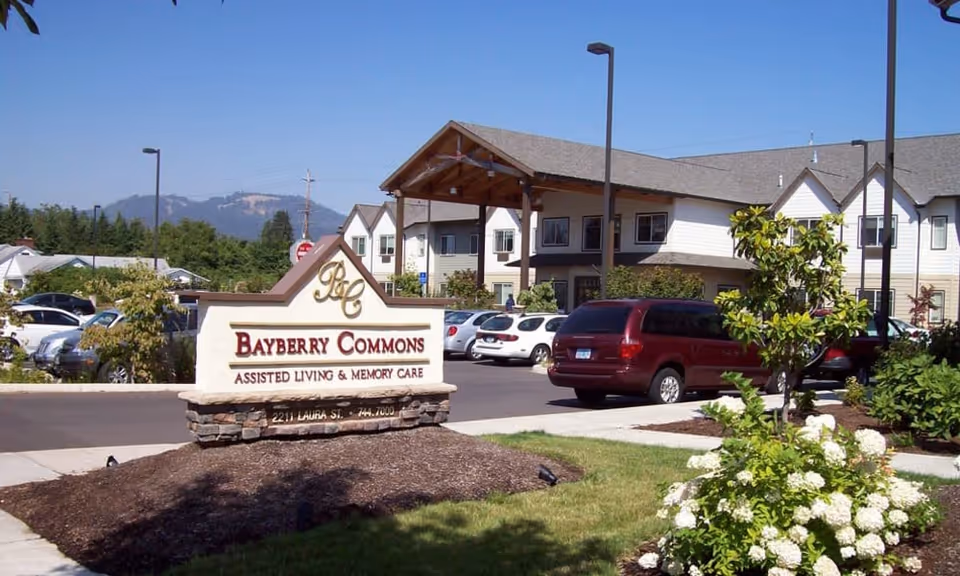 Exterior view of Bayberry Commons assisted living and memory care facility with a large sign in the foreground, a parking lot with several cars, landscaped greenery, and a two-story building under a clear blue sky.