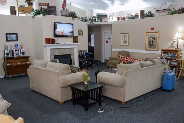 A cozy living room area in Ashley Manor Memory Care featuring two beige sofas arranged around a small black coffee table with a plant on it. A flat-screen TV is mounted above a fireplace on the far wall. The room has carpeted flooring, framed artwork on the walls, a wheelchair near the fireplace, and various decorative plants and items on a ledge above the walls. There is also a wooden dresser with a stereo system and a bookshelf with books and a lamp.