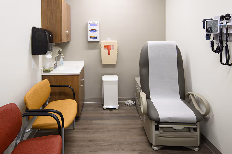 Small medical exam room with a paper-covered chair, sink, cabinets, and waiting chairs.