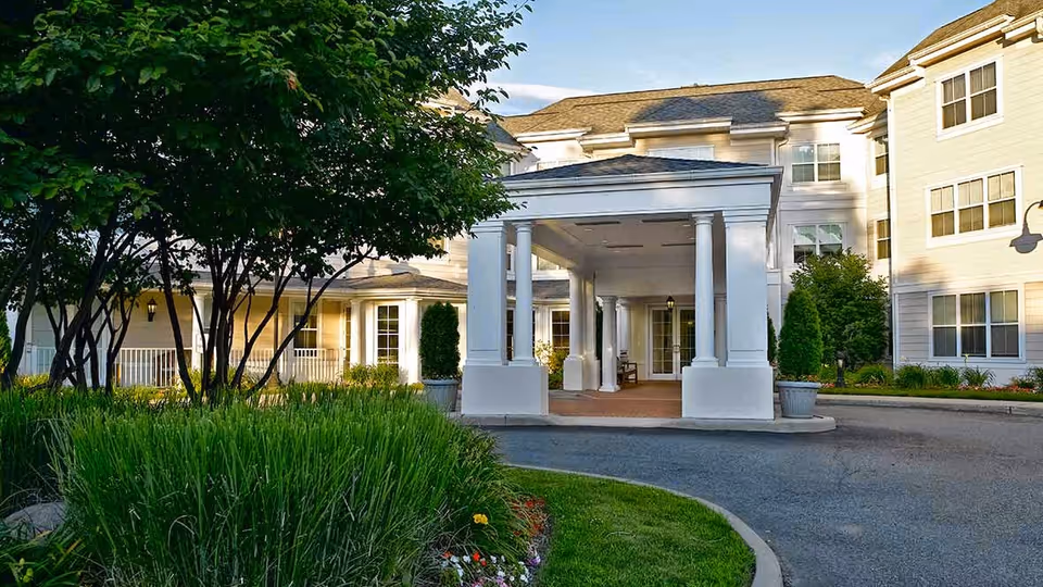 Front entrance/porte-cochere of a multi-story senior living building with white columns, landscaped lawns and a circular driveway.