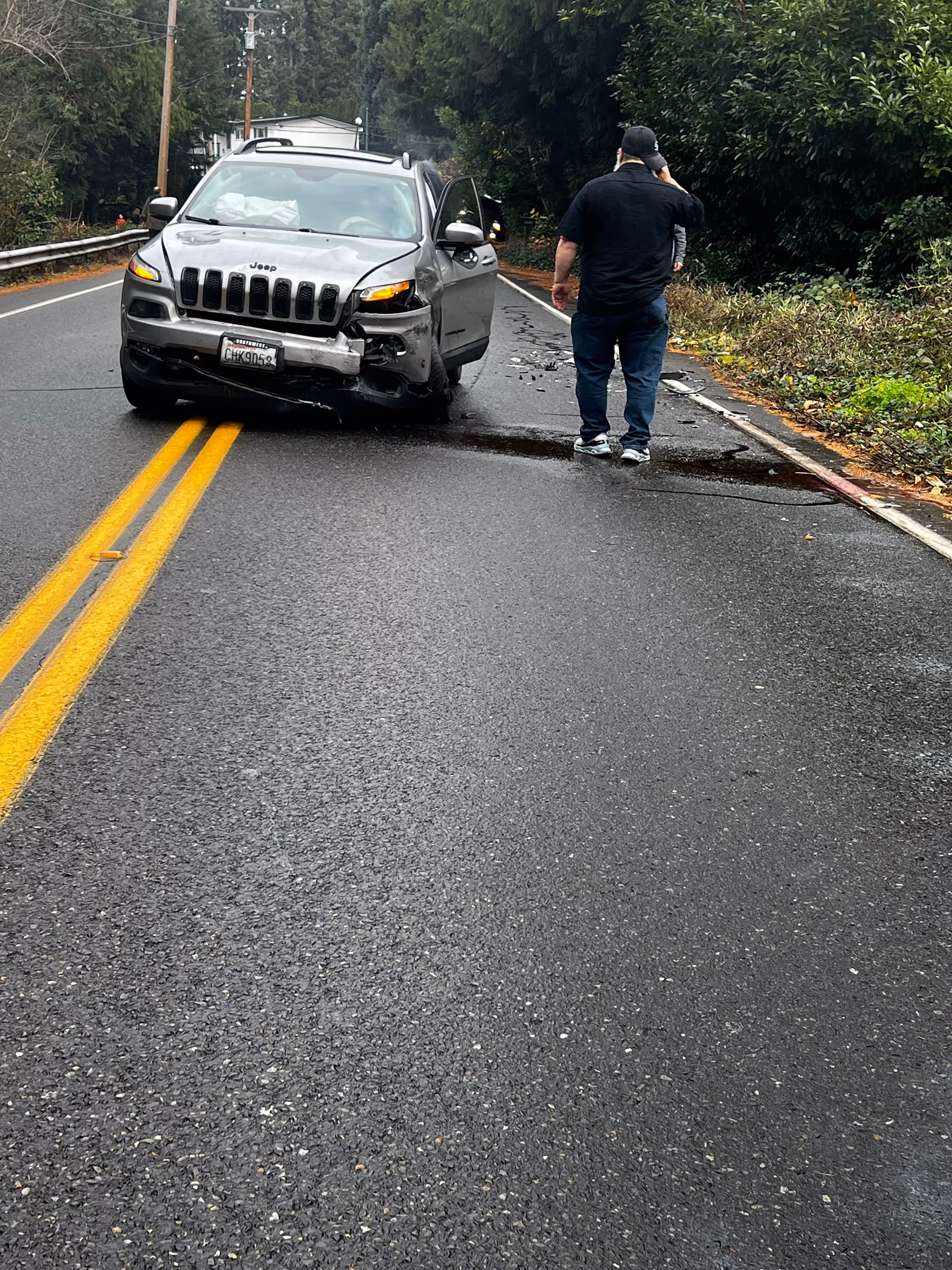 A silver Jeep with front-end damage on a wet road; a man stands on the shoulder.