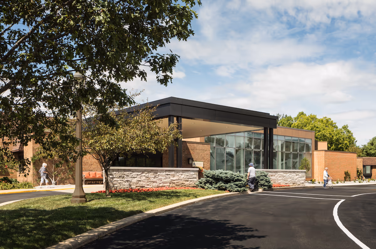Front entrance of a brick senior living building with a covered porte-cochere, landscaping, and a few people walking nearby.