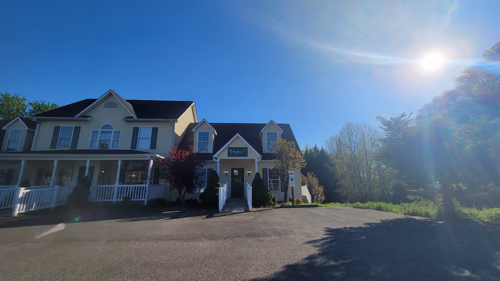 Two-story beige senior living building with white porch and surrounding trees.