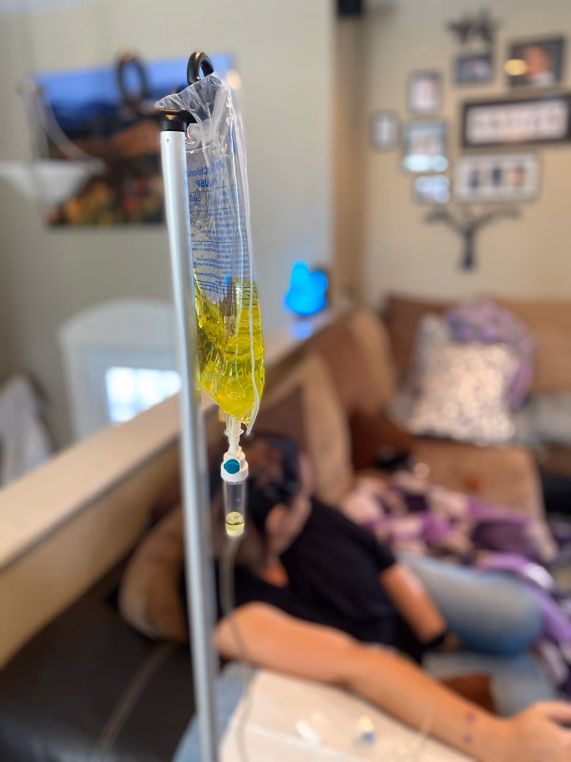 IV drip bag with yellow solution hangs beside a sofa in a living area.