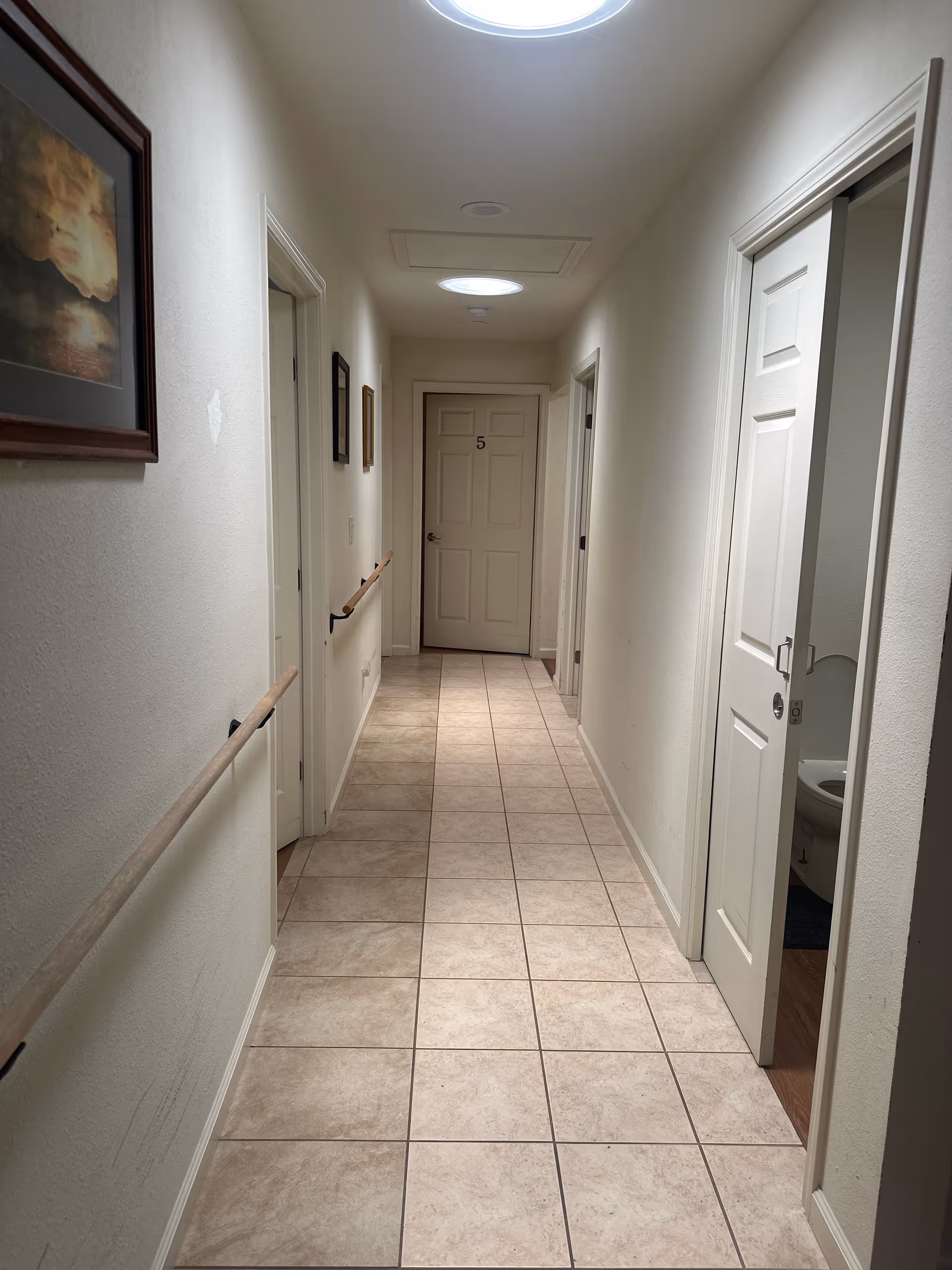 Long beige hallway in a senior living facility with doors, railings, and tiled floor.