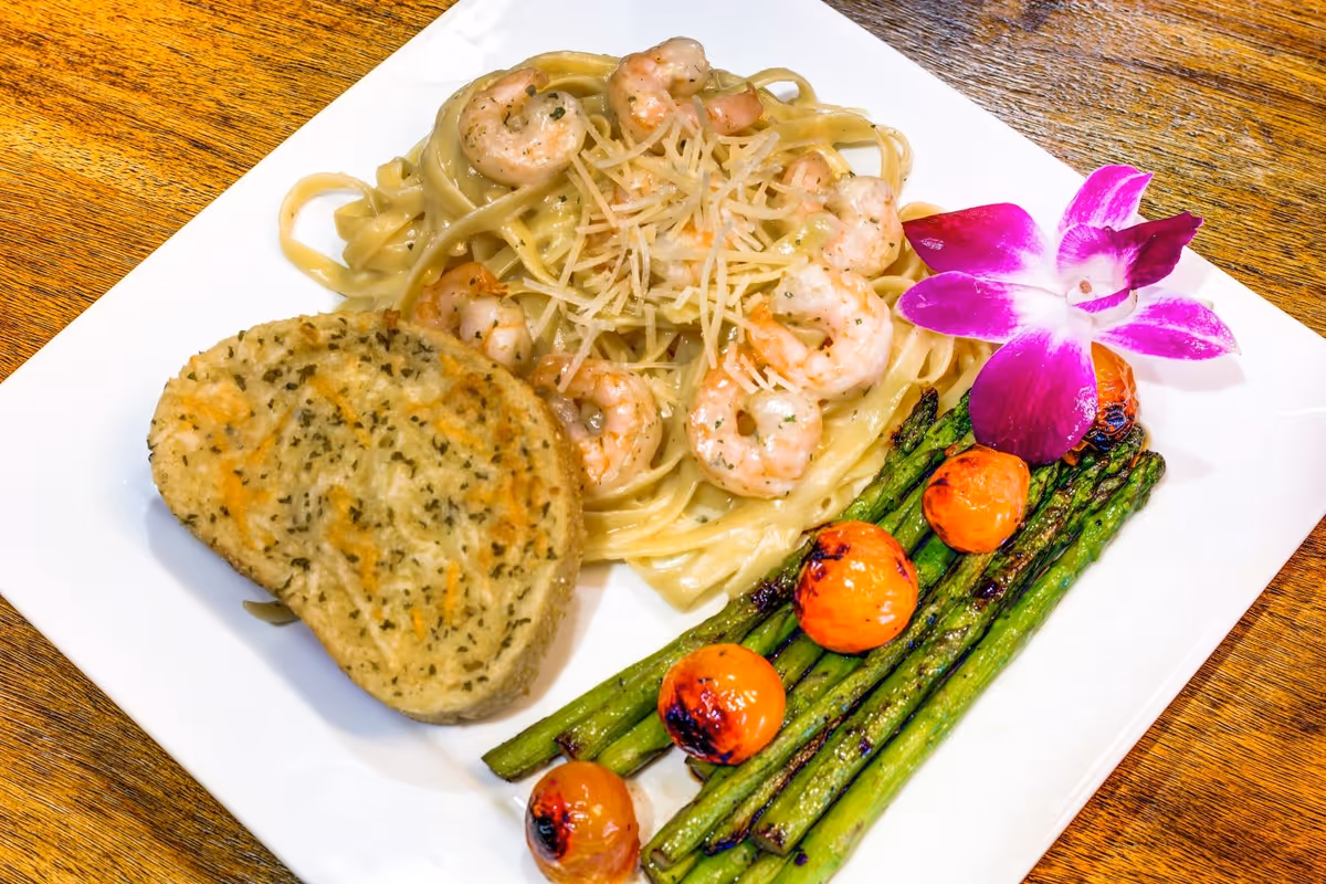 A white plate with shrimp fettuccine, garlic bread, grilled asparagus and cherry tomatoes garnished with a purple orchid on a wooden table.