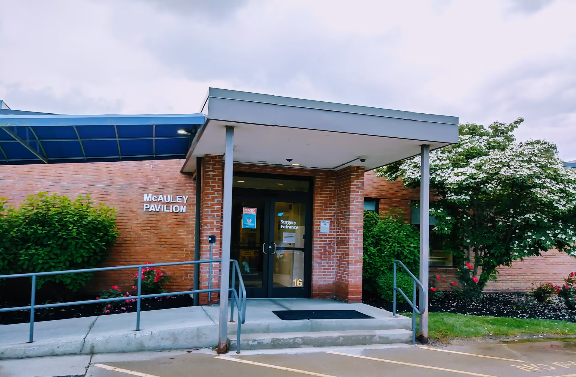 Brick hospital entrance with a blue canopy, glass doors, and surrounding greenery.