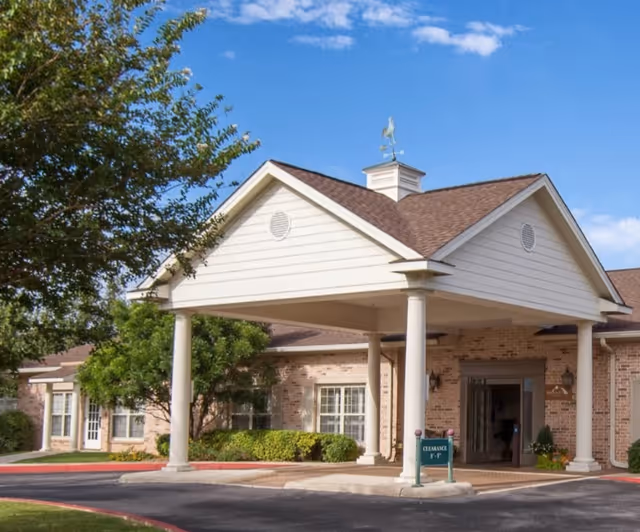 Entrance of a brick senior living facility with a covered portico supported by columns and surrounding landscaping.