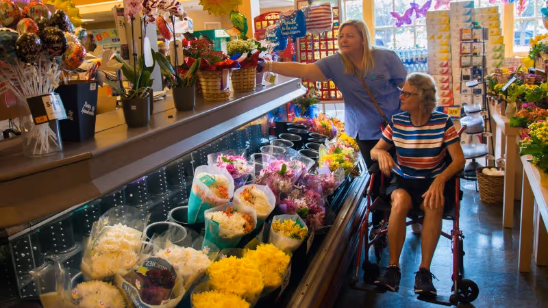 Staff member in scrubs assists a woman in a wheelchair at a flower counter.
