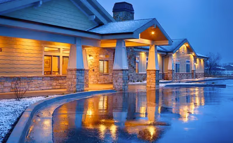 Evening exterior view of Seasons Assisted Living with illuminated entrance and curved water feature.