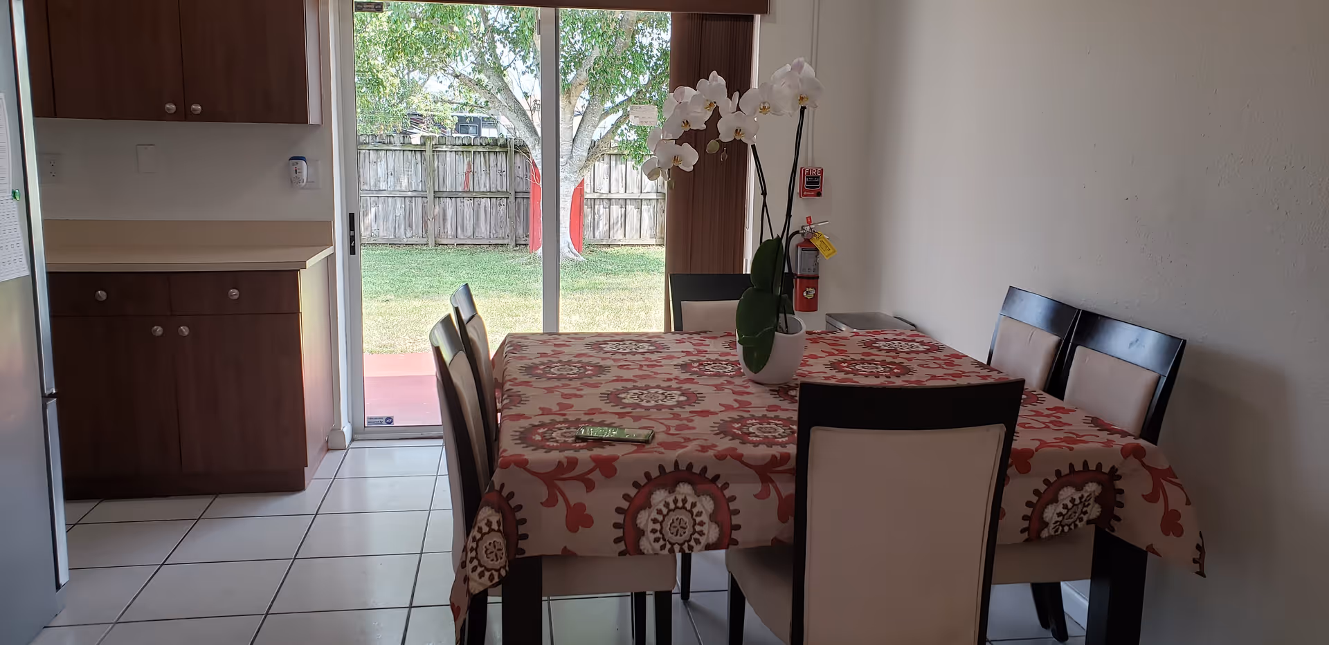 Dining area with a rectangular table covered with a patterned tablecloth and six chairs around it. A white orchid plant in a pot is placed on the table. The room has tiled flooring and wooden cabinets on the left side. A sliding glass door at the back opens to a fenced backyard with green grass and a tree.