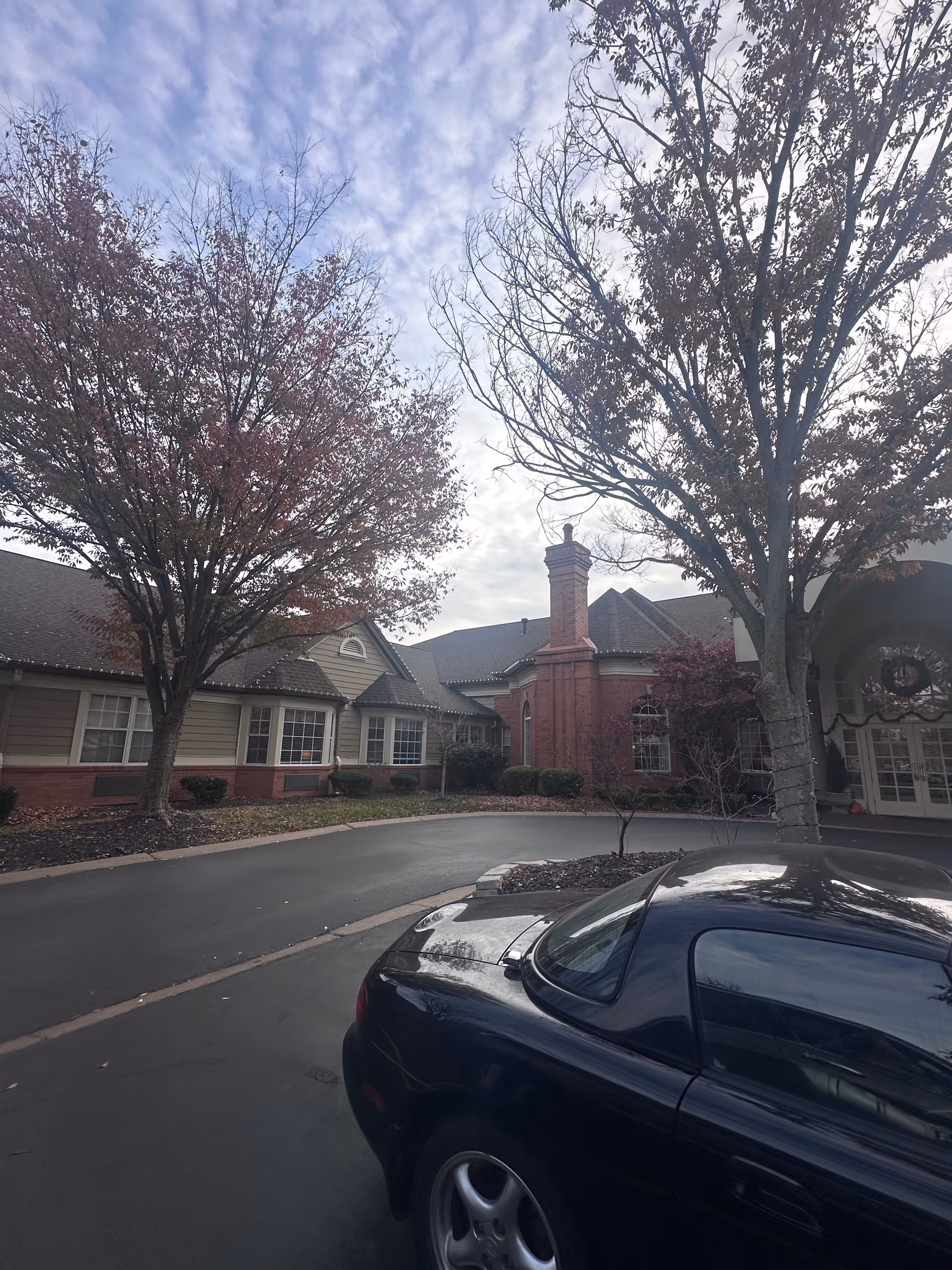 Front exterior of a brick senior living facility with a circular driveway, bare trees, and a parked black car under a cloudy sky.